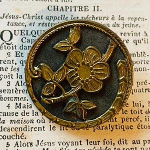 May include: Antique brass button with a floral design. The button features a raised flower with detailed petals and leaves, set against a dark background. The button has a decorative border and is set on a vintage book page.