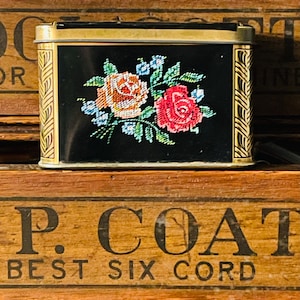 May include: A rectangular black metal tin featuring a central floral design of orange, red, and blue roses with green leaves. The tin has a gold-colored lid and gold-colored side panels decorated with a repeating vertical leaf pattern.