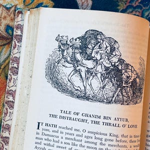 May include: Open book with a black and white illustration of people riding camels. The text reads "Tale of Ghanim Bin Ayyub, The Distraught, The Thrall O' Love." The book's pages are aged, and the cover has a floral design.