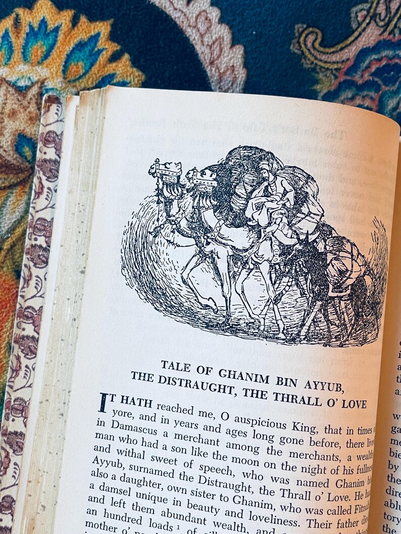 May include: Open book with a black and white illustration of people riding camels. The text reads "Tale of Ghanim Bin Ayyub, The Distraught, The Thrall O' Love." The book's pages are aged, and the cover has a floral design.