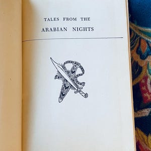 May include: Open book with the title "TALES FROM THE ARABIAN NIGHTS" printed in black. Below the title is a black and white illustration of a dagger and scabbard. The book's pages are off-white.