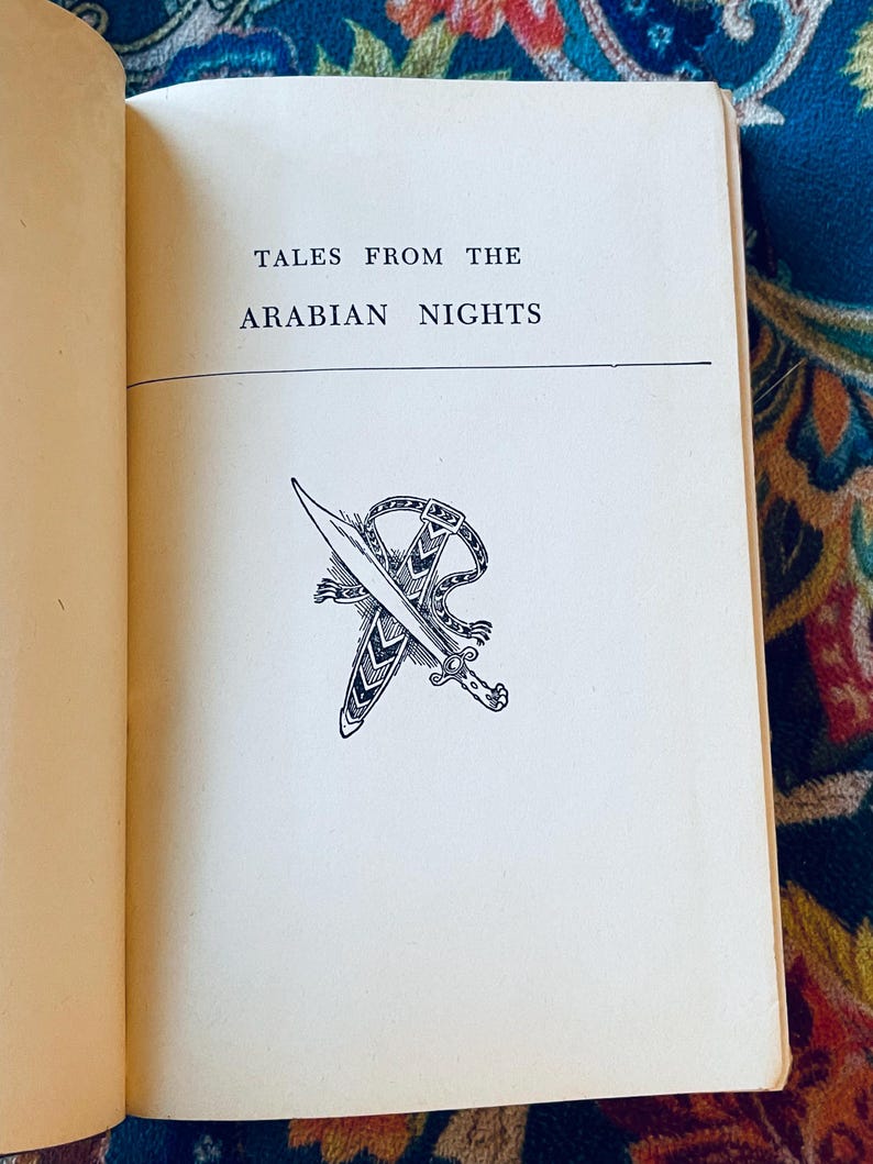 May include: Open book with the title "TALES FROM THE ARABIAN NIGHTS" printed in black. Below the title is a black and white illustration of a dagger and scabbard. The book's pages are off-white.
