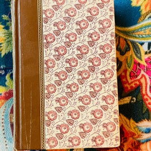 May include: A hardcover book with a brown spine and a floral pattern cover in cream and pink. The cover design features repeating pink flowers with brown stems and leaves. The book is closed and resting on a colorful patterned surface.