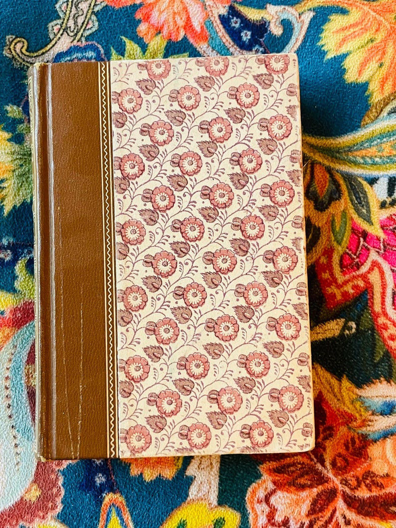 May include: A hardcover book with a brown spine and a floral pattern cover in cream and pink. The cover design features repeating pink flowers with brown stems and leaves. The book is closed and resting on a colorful patterned surface.