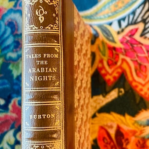 May include: A vintage hardcover book titled "Tales From The Arabian Nights" by Burton. The book has a brown leather cover with gold lettering and decorative accents. The spine is visible, showcasing the title and author's name.