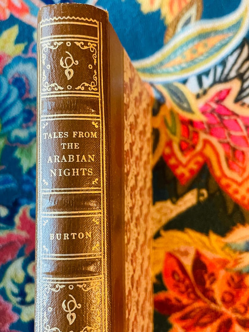 May include: A vintage hardcover book titled "Tales From The Arabian Nights" by Burton. The book has a brown leather cover with gold lettering and decorative accents. The spine is visible, showcasing the title and author's name.