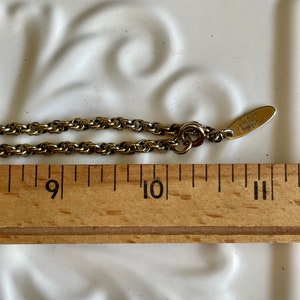 K&ouml;nnte beinhalten: Eine goldfarbene Halskette mit einem kleinen Anh&auml;nger mit der Aufschrift "AVON". Die Kette wird mit einem Holzlineal gemessen, das L&auml;ngen zwischen 23 und 28 cm anzeigt. Der Hintergrund ist eine wei&szlig;e Oberfl&auml;che mit einem dekorativen Muster.