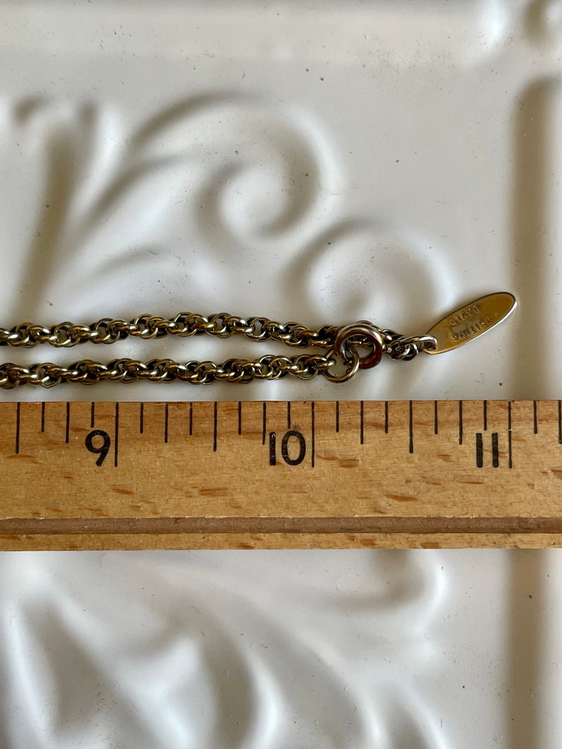 K&ouml;nnte beinhalten: Eine goldfarbene Halskette mit einem kleinen Anh&auml;nger mit der Aufschrift "AVON". Die Kette wird mit einem Holzlineal gemessen, das L&auml;ngen zwischen 23 und 28 cm anzeigt. Der Hintergrund ist eine wei&szlig;e Oberfl&auml;che mit einem dekorativen Muster.