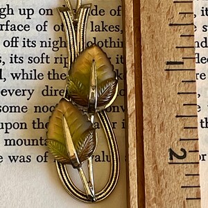 K&ouml;nnte beinhalten: Eine Vintage-Halskette mit goldfarbenem Anh&auml;nger und Blattdesign. Der Anh&auml;nger hat zwei detaillierte Bl&auml;tter in Gr&uuml;n- und Braunt&ouml;nen mit einem silberfarbenen Stiel. Die Halskette wird mit einem Holzlineal pr&auml;sentiert.