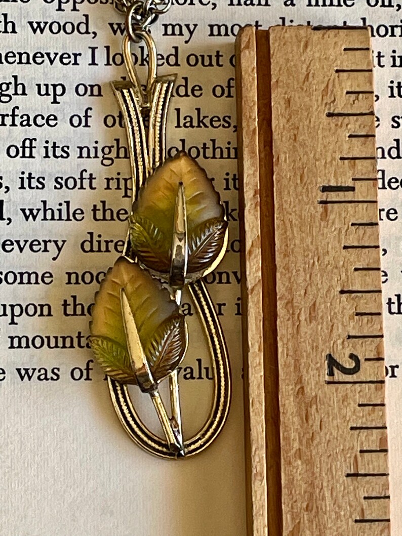 K&ouml;nnte beinhalten: Eine Vintage-Halskette mit goldfarbenem Anh&auml;nger und Blattdesign. Der Anh&auml;nger hat zwei detaillierte Bl&auml;tter in Gr&uuml;n- und Braunt&ouml;nen mit einem silberfarbenen Stiel. Die Halskette wird mit einem Holzlineal pr&auml;sentiert.