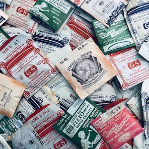 May include: A collection of vintage watch crystal packets in various sizes and colors. The packets feature text such as "G-S Flexo Crystals" and "Super Quality Watch Crystals." Some packets have measurements like "41.9mm" and "Size L 56."