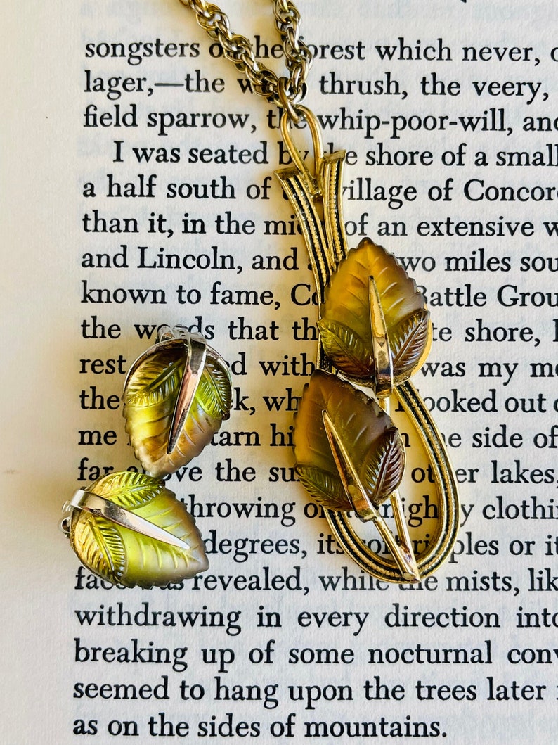 K&ouml;nnte beinhalten: Eine Vintage-Halskette in Goldoptik und ein passendes Ohrring-Set. Der Anh&auml;nger und die Ohrringe haben blattf&ouml;rmige Akzente in Bernstein- und Gr&uuml;nt&ouml;nen. Der Schmuck wird auf einer Seite mit gedrucktem Text pr&auml;sentiert.