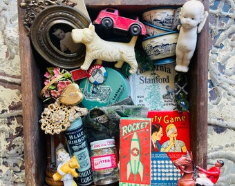 May include: A wooden box filled with vintage toys and trinkets, including a red toy car, a white porcelain dog, a porcelain angel figurine, a small framed photo, a blue and white teacup, a small tin with a picture of John F. Kennedy, a small tin with a picture of a boy, a small tin with the words "Blue Ribbon", a small jar of glitter, a small box of chalk, a small box of "Rocket" firecrackers, a small box of "Party Game" for adults, and a small brown and white horse figurine with a rider.