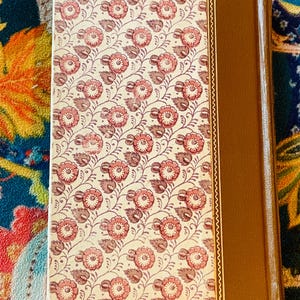 May include: A vintage book with a floral pattern on the cover. The cover features a cream background with a repeating pattern of pink flowers and brown leaves. The spine is a solid brown color.