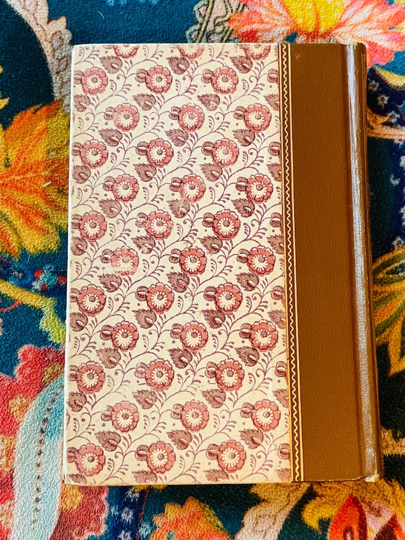 May include: A vintage book with a floral pattern on the cover. The cover features a cream background with a repeating pattern of pink flowers and brown leaves. The spine is a solid brown color.