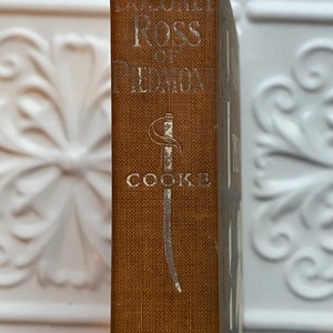 May include: A brown book with the title "Colonel Ross of Piedmont" and the author "Cooke" printed on the spine. A silver sword design is featured on the spine.