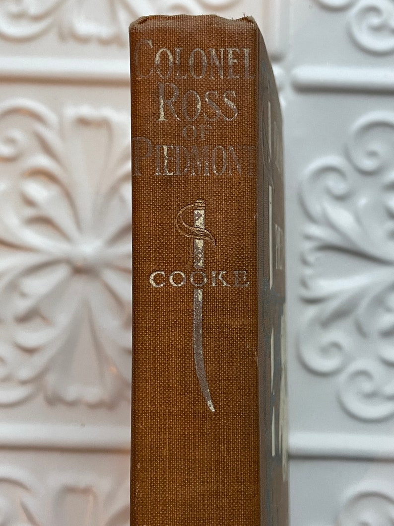 May include: A brown book with the title "Colonel Ross of Piedmont" and the author "Cooke" printed on the spine. A silver sword design is featured on the spine.
