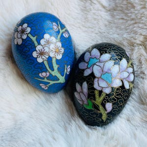 May include: Two decorative, egg-shaped ornaments. One is blue with white floral designs and gold accents. The other is black with a floral pattern in shades of purple, blue, and gold. Both have a glossy finish and are set against a white, furry background.