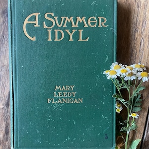 May include: A vintage green hardcover book titled A Summer Idyl by Mary Leedy Flanigan with gold embossed lettering on the front cover, accompanied by a small sprig of white and yellow chamomile flowers with green leaves and stems.