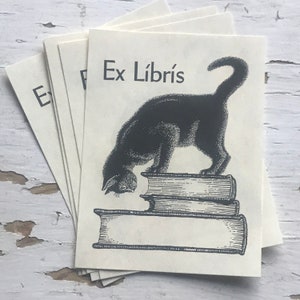 May include: A black cat with a white chest is looking at a stack of books. The books are stacked on top of each other and have a vintage look. The text "Ex Libris" is printed on the top of the book stack.