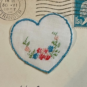 May include: A heart-shaped embroidered patch with a blue stitched border. The white fabric features a floral design in pink, red, and blue. The patch is on a vintage envelope with a stamp and postmark.