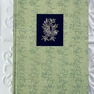May include: A hardcover book with a light green floral pattern. A dark blue square in the center features a gold-colored letter "D" surrounded by ornate floral designs. The book's cover has a textured appearance.