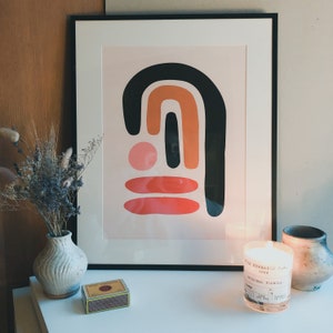 May include: A framed abstract art print with black, orange, and red shapes. A lit candle, a small vase with dried flowers, a matchbox, and a ceramic vase are on a white surface. The candle has a label that reads "Vila Hermanos S.A. 1884 ORIGINAL FORMULA".