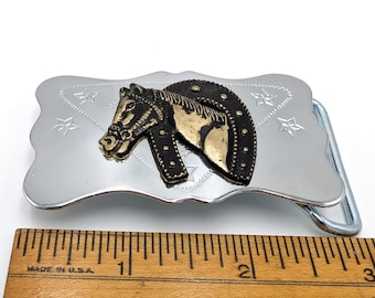 May include: Silver belt buckle with a gold and black horse head design. The horse head is wearing a horseshoe. The buckle is decorated with stars.