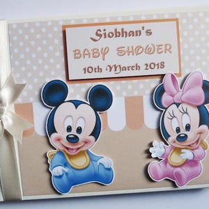 May include: A baby shower memory book with a cream-colored ribbon and bow on the left side. The cover features Mickey and Minnie Mouse cutouts, and a sign that reads "Siobhan's Baby Shower 10th March 2018."