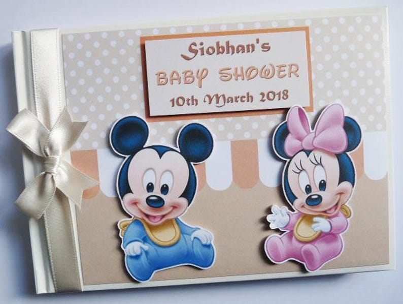 May include: A baby shower memory book with a cream-colored ribbon and bow on the left side. The cover features Mickey and Minnie Mouse cutouts, and a sign that reads "Siobhan's Baby Shower 10th March 2018."