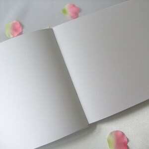 Puede incluir: Un cuaderno de bocetos abierto con p&aacute;ginas blancas en blanco. El libro est&aacute; sobre una superficie blanca, con p&eacute;talos de flores rosas y verdes esparcidos. Las p&aacute;ginas est&aacute;n limpias y listas para dibujar o escribir.