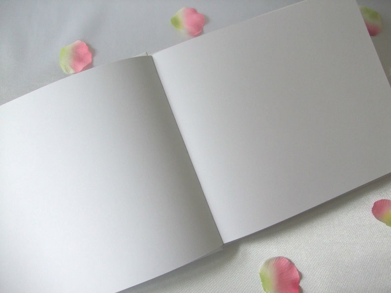 Puede incluir: Un cuaderno de bocetos abierto con p&aacute;ginas blancas en blanco. El libro est&aacute; sobre una superficie blanca, con p&eacute;talos de flores rosas y verdes esparcidos. Las p&aacute;ginas est&aacute;n limpias y listas para dibujar o escribir.