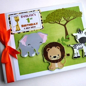 Puede incluir: Un &aacute;lbum de recortes de cumplea&ntilde;os personalizado con tem&aacute;tica de safari. La portada presenta una jirafa, un elefante, un le&oacute;n y una cebra sobre un fondo verde. El texto "Evelyn's 1st Birthday 5 July 2014" est&aacute; en la portada. Una cinta naranja est&aacute; atada en el lado izquierdo.