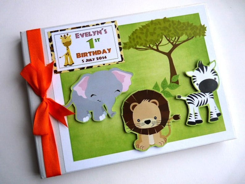 Puede incluir: Un &aacute;lbum de recortes de cumplea&ntilde;os personalizado con tem&aacute;tica de safari. La portada presenta una jirafa, un elefante, un le&oacute;n y una cebra sobre un fondo verde. El texto "Evelyn's 1st Birthday 5 July 2014" est&aacute; en la portada. Una cinta naranja est&aacute; atada en el lado izquierdo.