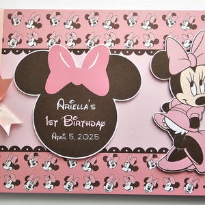 Puede incluir: Un álbum de recortes rosa y marrón con un tema de Minnie Mouse. La portada presenta una gran cabeza marrón de Minnie Mouse con un lazo rosa y el texto "Ariella's 1st Birthday April 5, 2025". También hay una figura más pequeña de Minnie Mouse en la portada.