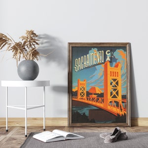 May include: A vintage-style poster featuring a stylised illustration of the Tower Bridge in Sacramento, California. The poster is framed in a dark wood frame and has a blue, orange, and yellow colour scheme. The text "Sacramento CA" is written in a stylised font at the top of the poster.