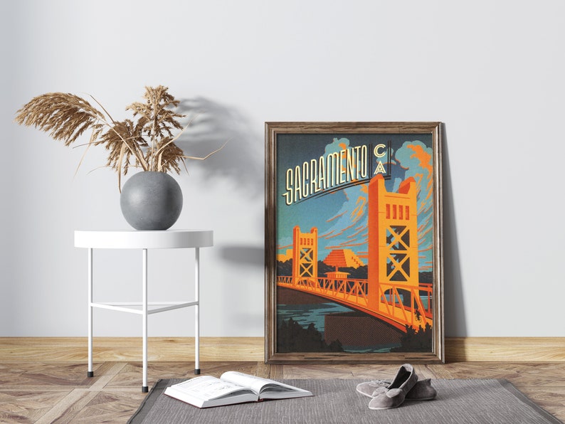May include: A vintage-style poster featuring a stylised illustration of the Tower Bridge in Sacramento, California. The poster is framed in a dark wood frame and has a blue, orange, and yellow colour scheme. The text "Sacramento CA" is written in a stylised font at the top of the poster.