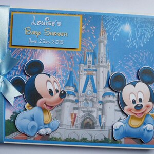 May include: A baby shower guest book featuring Mickey Mouse illustrations. The cover has a blue background with a castle and fireworks. The text reads "Louise's Baby Shower June 23rd 2018". A light blue ribbon is tied on the left side.