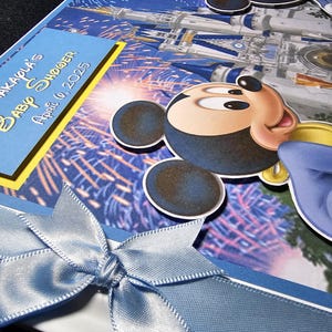 May include: A baby shower invitation featuring Mickey Mouse and a castle design. The invitation has a blue satin ribbon bow and text that reads "AKAPLA'S Baby Shower April 6, 2025". The background is a blue and white fireworks display.