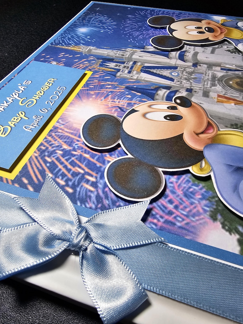 May include: A baby shower invitation featuring Mickey Mouse and a castle design. The invitation has a blue satin ribbon bow and text that reads "AKAPLA'S Baby Shower April 6, 2025". The background is a blue and white fireworks display.