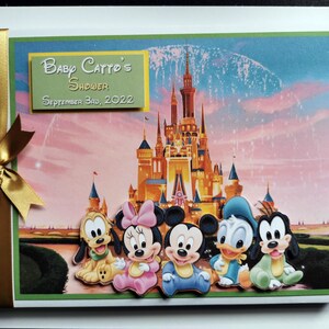 May include: A baby shower scrapbook with a gold ribbon and bow. The cover features a Disney castle illustration and character cutouts of Pluto, Minnie Mouse, Mickey Mouse, Donald Duck, and Goofy. The text reads "Baby Catto's Shower September 3rd, 2022."
