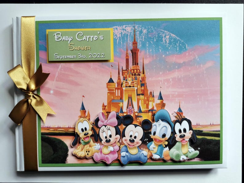 May include: A baby shower scrapbook with a gold ribbon and bow. The cover features a Disney castle illustration and character cutouts of Pluto, Minnie Mouse, Mickey Mouse, Donald Duck, and Goofy. The text reads "Baby Catto's Shower September 3rd, 2022."
