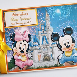 May include: A baby shower scrapbook featuring Minnie and Mickey Mouse. The cover displays a vibrant illustration of Cinderella's castle with fireworks. The text "Georgina's Baby Shower 10th February 2020" is printed on a gold label. A yellow ribbon is tied on the spine.