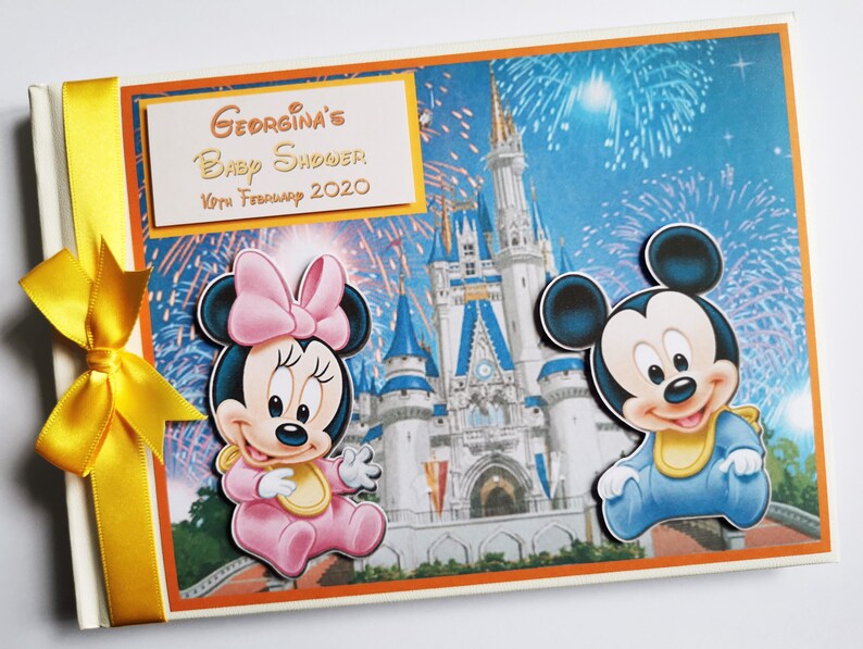 May include: A baby shower scrapbook featuring Minnie and Mickey Mouse. The cover displays a vibrant illustration of Cinderella's castle with fireworks. The text "Georgina's Baby Shower 10th February 2020" is printed on a gold label. A yellow ribbon is tied on the spine.