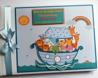 Op de afbeelding: Een blauw en wit gastenboek met een regenboog, zon en een Noach's Ark-thema. Het boek is versierd met een blauw lint en de tekst "Harry George Love Christening Guest Book 8th September 2018".