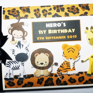 Puede incluir: Un &aacute;lbum de recortes blanco con una cinta negra, con recortes de animales de un mono, cebra, le&oacute;n, tigre y jirafa. La portada incluye el texto "HERO'S 1ST BIRTHDAY 6TH SEPTEMBER 2019" con un borde de estampado de leopardo.