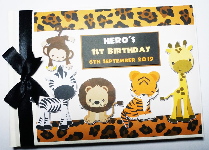 Puede incluir: Un &aacute;lbum de recortes blanco con una cinta negra, con recortes de animales de un mono, cebra, le&oacute;n, tigre y jirafa. La portada incluye el texto "HERO'S 1ST BIRTHDAY 6TH SEPTEMBER 2019" con un borde de estampado de leopardo.