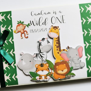 Puede incluir: Un &aacute;lbum de fotos blanco con una cinta verde e ilustraciones con tem&aacute;tica de jungla. La portada dice "CamCam is a Wild One" con una fecha. El &aacute;lbum presenta animales de dibujos animados, incluyendo un mono, una cebra, una jirafa, un elefante, un tigre, un rinoceronte y un le&oacute;n.