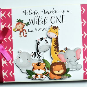 Puede incluir: &Aacute;lbum de fotos de beb&eacute; personalizado con lazo rosa. La portada presenta el texto "Melody Aurelia is a Wild One" e ilustraciones de animales de la jungla, incluyendo un mono, una jirafa, una cebra, un tigre, un le&oacute;n, un rinoceronte y un elefante.