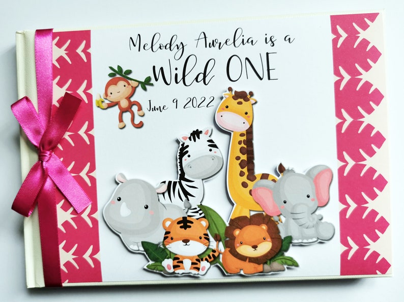 Puede incluir: &Aacute;lbum de fotos de beb&eacute; personalizado con lazo rosa. La portada presenta el texto "Melody Aurelia is a Wild One" e ilustraciones de animales de la jungla, incluyendo un mono, una jirafa, una cebra, un tigre, un le&oacute;n, un rinoceronte y un elefante.